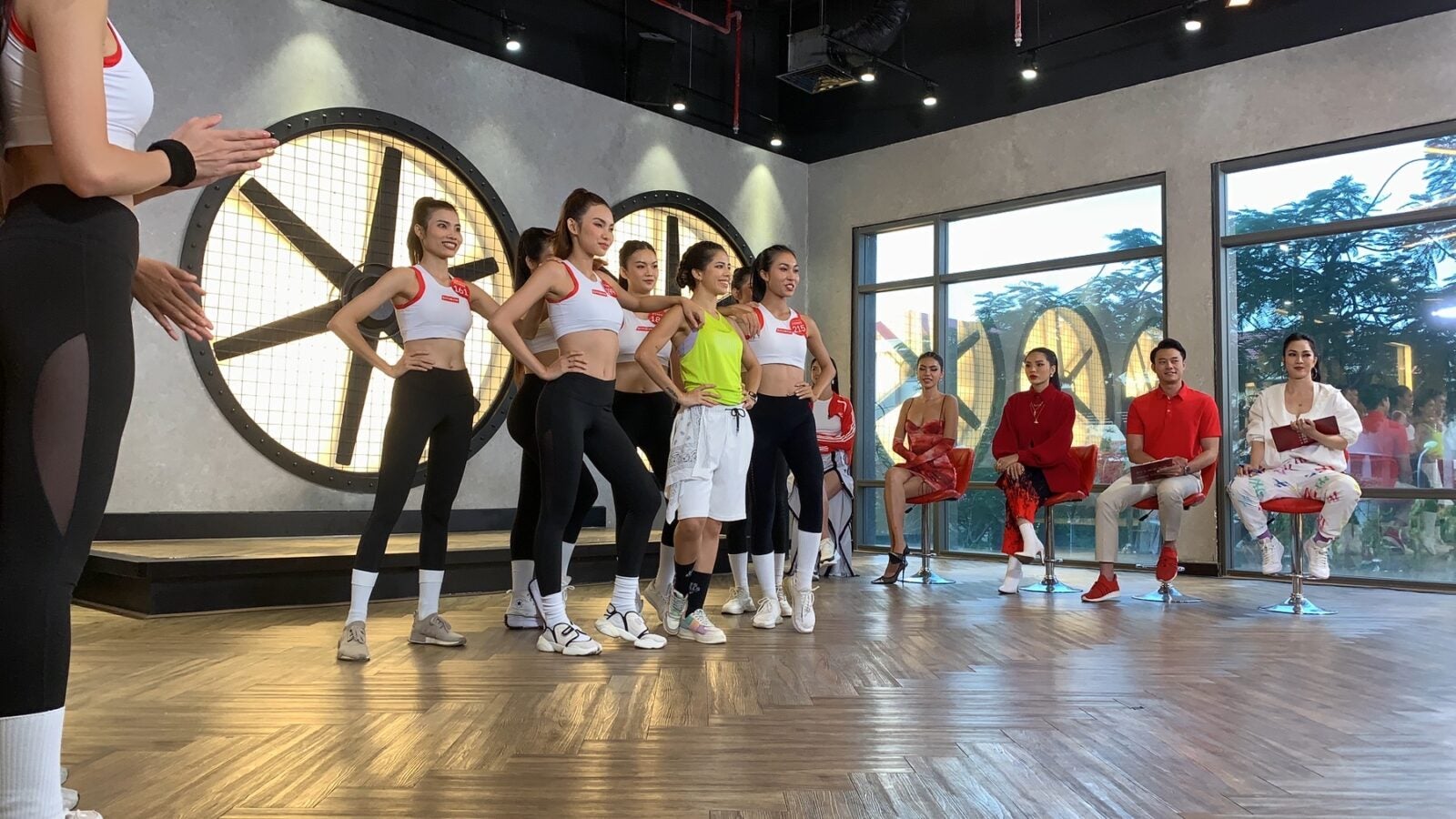 A group of fitness enthusiasts stand posing together wearing white tank tops and black pants while one woman stands in the middle wearing a yellow tank top and white pants. Four non-participants are sitting at the side while watching intently.