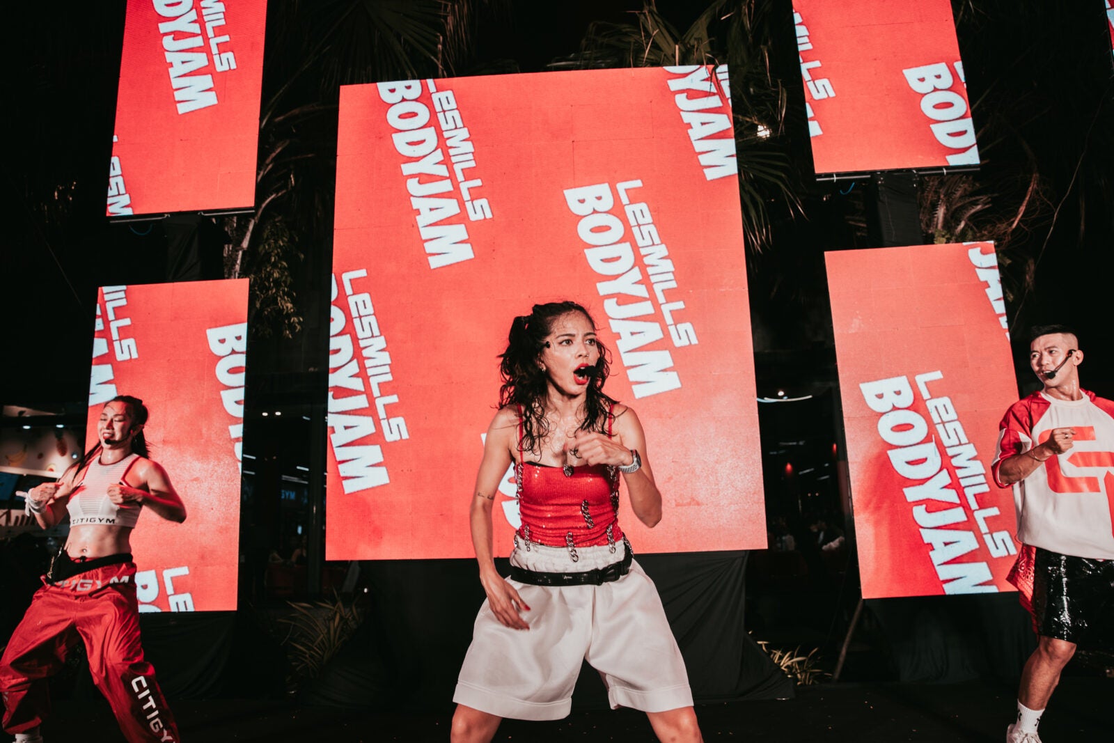 A Young Energised Asian Woman In White Shorts And Red Tank Stands In The Middle Of A Stage While Talking Through A Microphone Attachment. Behind Her Are Two Other Fitness Instructors Mirroring Her And The Company'S Branding &Quot;Les Mills Bodyjam&Quot; Printed In The Background.