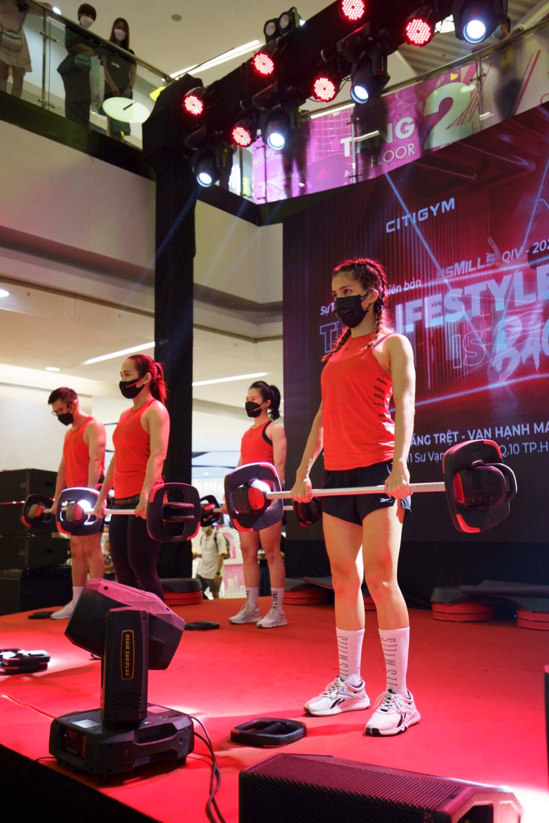 Four fitness instructors, three woman and one man who are holding onto weights and standing on a stage in a mall as people from the upper levels look on.