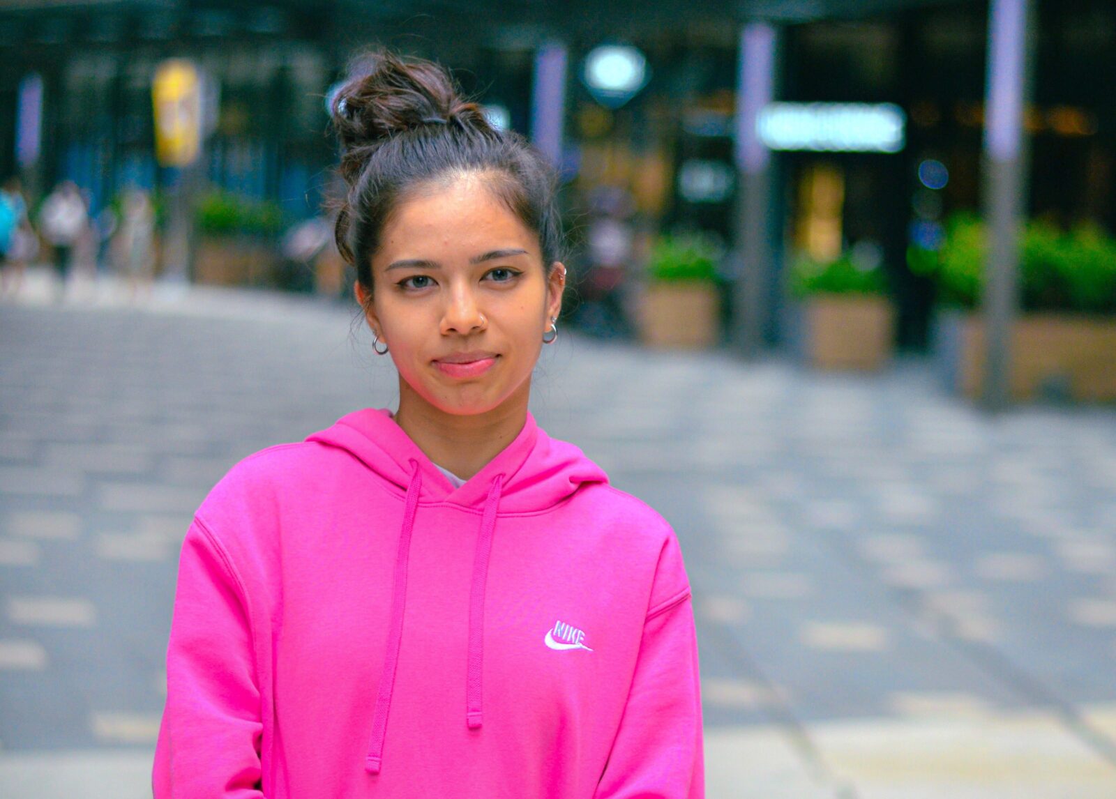 A young woman wearing a hot pink hoodie with her hair tied in a messy bun poses at the side walkway in Lalaport.