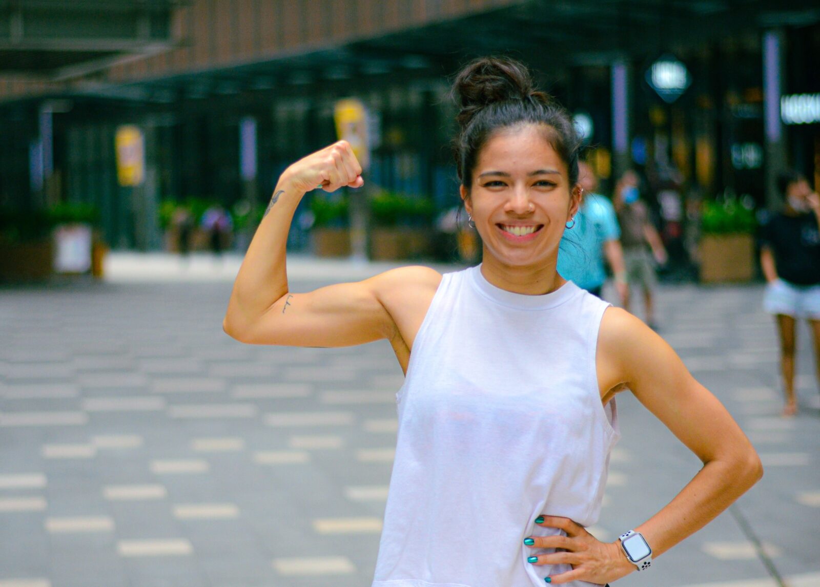 A young Asian woman wearing her hair in a bun and a white loose tank top posing to show her biceps while smiling