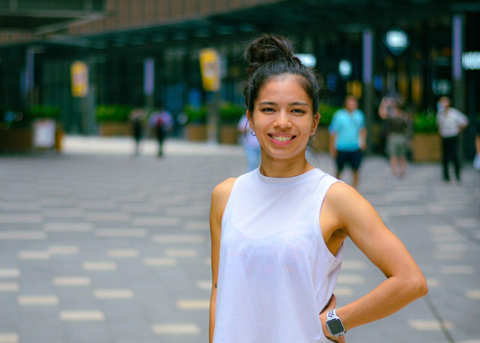 A young Asian woman wearing her hair in a bun and a white loose tank top posing while smiling