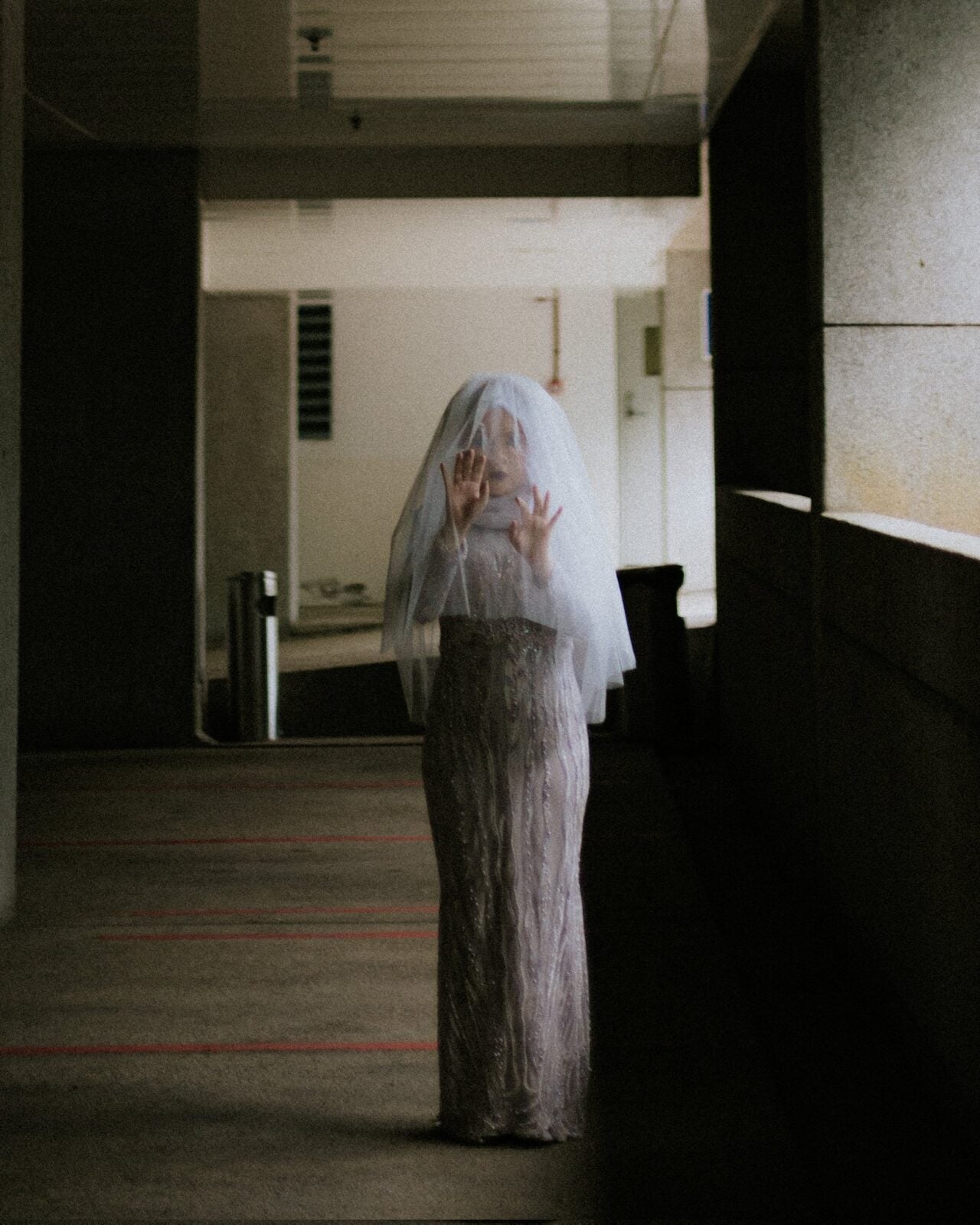 A woman wearing a white dress and hijab covered in a white veil stands creepily in a dark hallway with her hands reaching out in front of her.