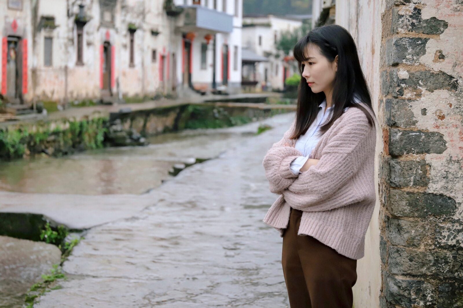 A young Asian woman wearing a beige cardigan, white shirt and brown pants stand with her hands crossed over each other while looking unhappily in the distance.