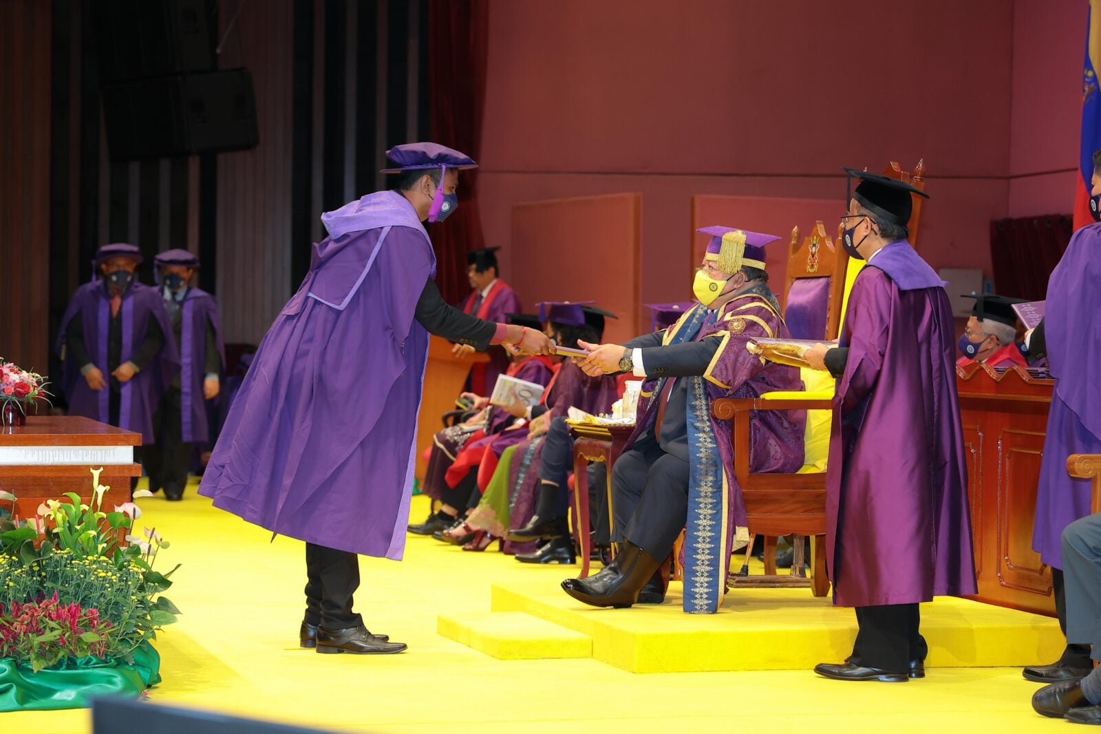 A graduation ceremony for Masters students where a man is being handed his certificate by another man who is sitting.