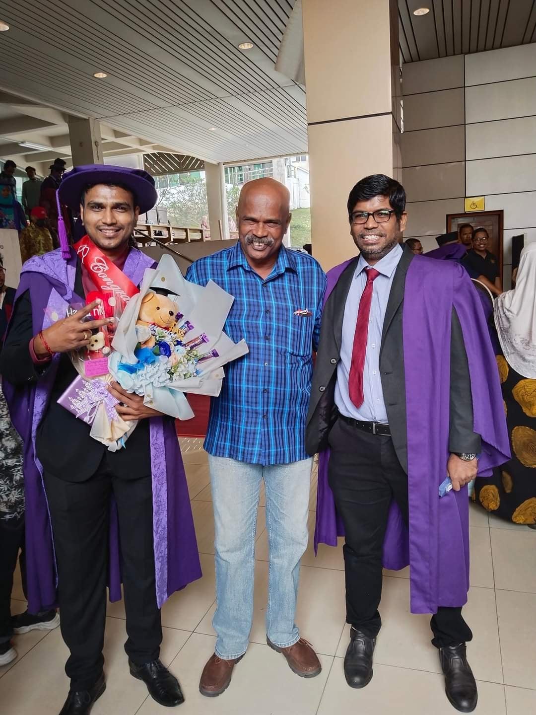 Three Indian men smiling, two of whom are wearing a purple graduation robe and one man holding a bouquet of chocolates and a teddybear.