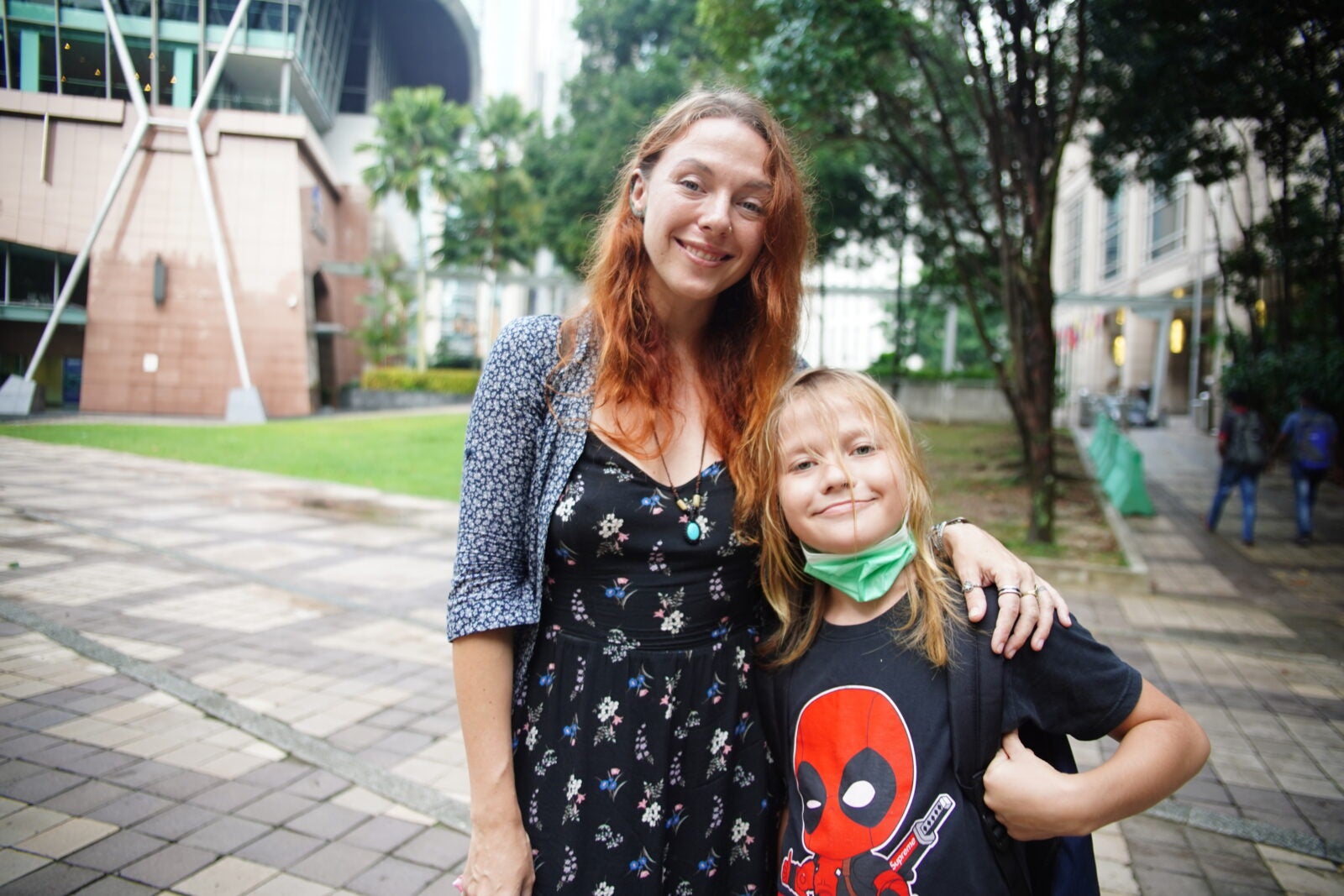 A ginger-haired Russian woman and her blonde son stand posing and smiling at a park
