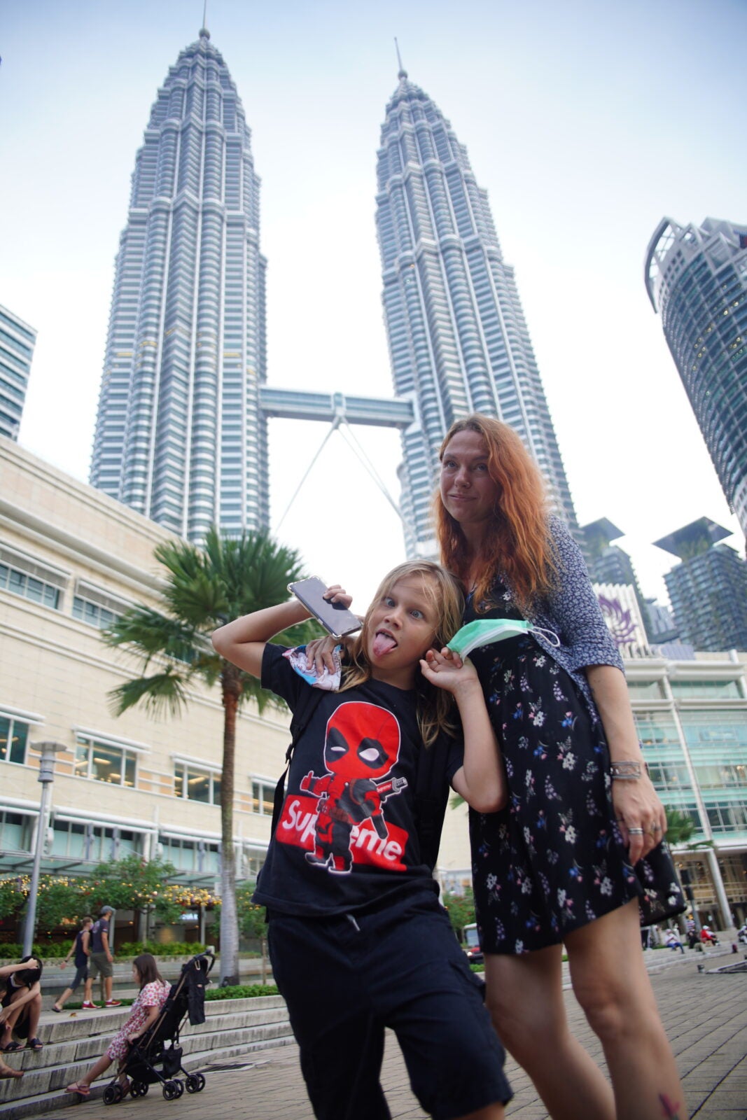 In fron of the Kuala Lumpur Twin Towers, a ginger-haired Russian woman wearing a grey cardigan over her dress stands beside her blonde son who is wearing a Deadpool shirt while making a silly face.