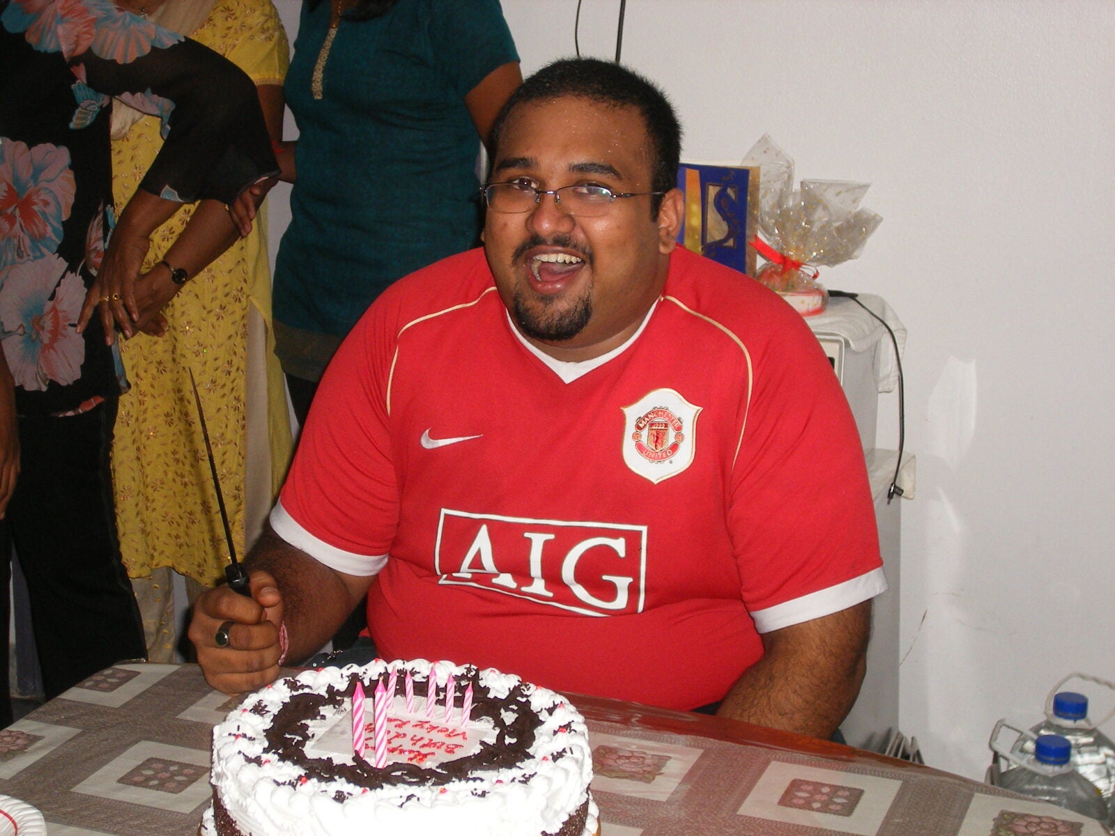 An Indian Man With Short Hair And Square Glasses Smile With A 12 Inch Cake In Front Of Him.