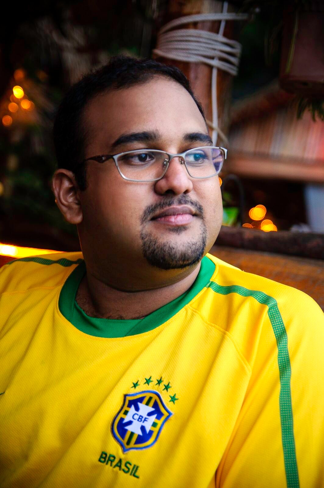 An Indian Bearded Man Wearing Glasses And Brazil'S Football Jersey Looking To His Left.