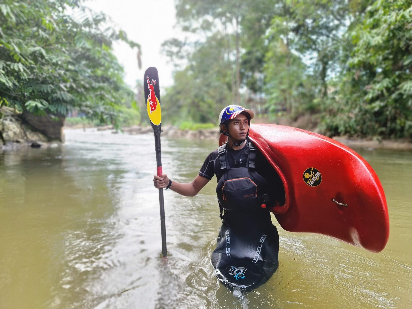 A Malay man stands in a knee-deep river. He holds a red kayak over his shoulders and its paddle in his right hand.
