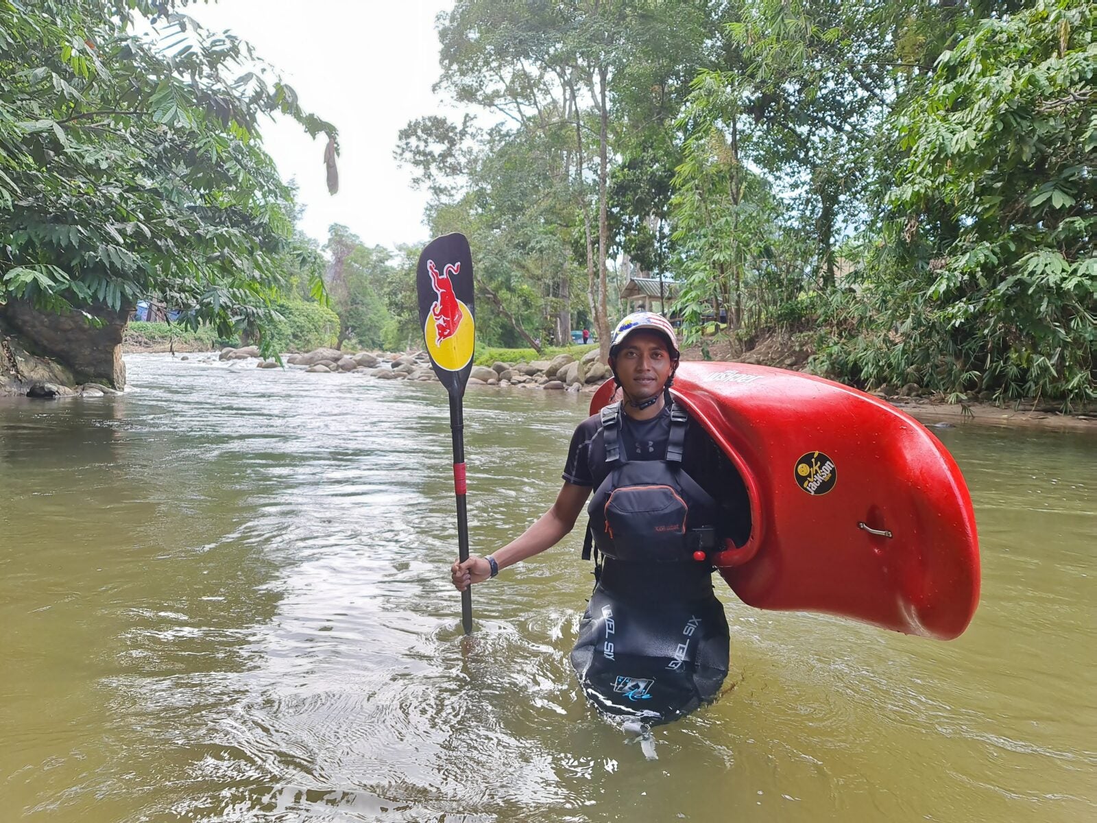 A Malay man stands and looks straight on while he is in a knee-deep river. He holds a red kayak over his shoulders and its paddle in his right hand.