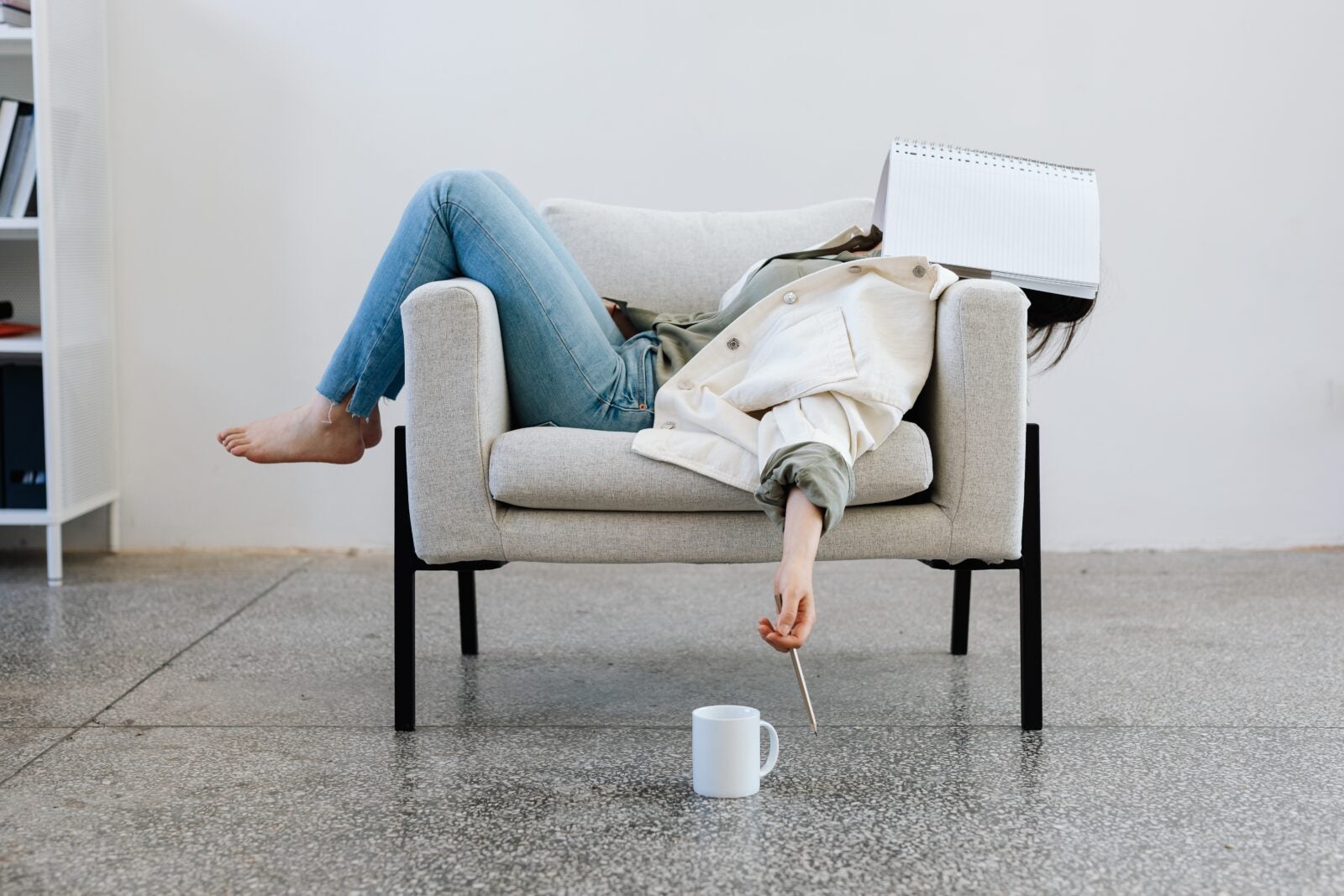 A woman laying on a one-seater sofa with her legs hanging off the armrest and a book covering her face. Her hands hang on the front of the sofa while holding a pencil and a cup is on the floor nearby.