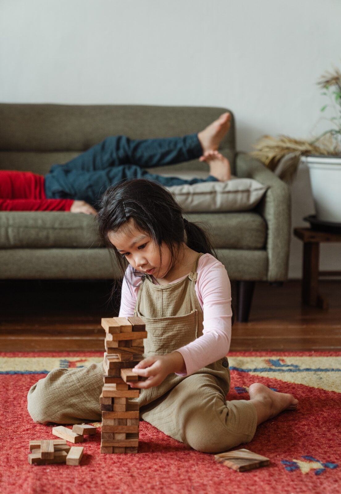 A young Asian girl playing with small wooden blocks that are stacked like Jenga while an adult rests behind her on a sofa.