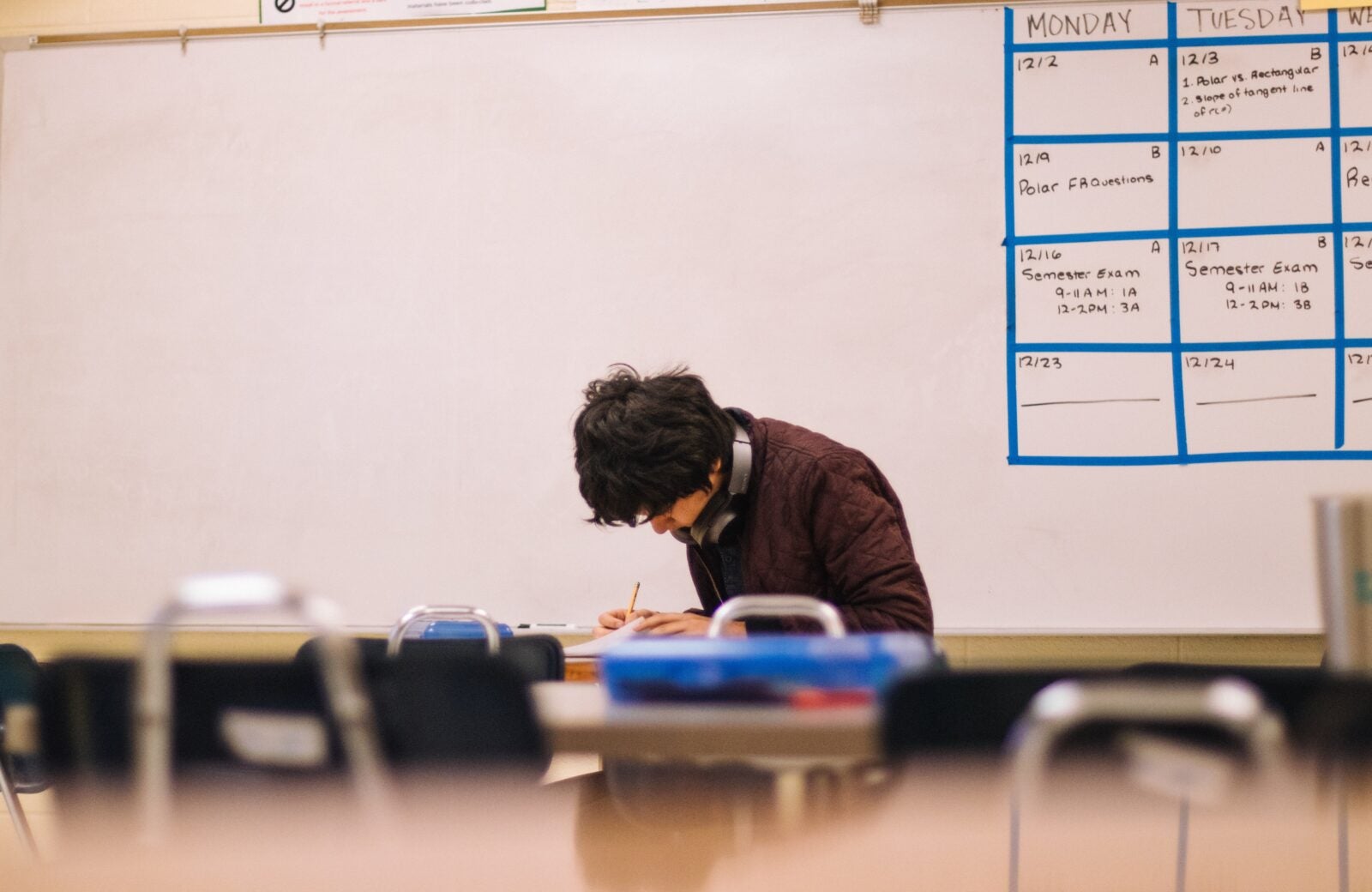 Young man wearing a brown sweater and resting his headphones on his neck sits alone in a classroom while looking down and writing on a notebook. 