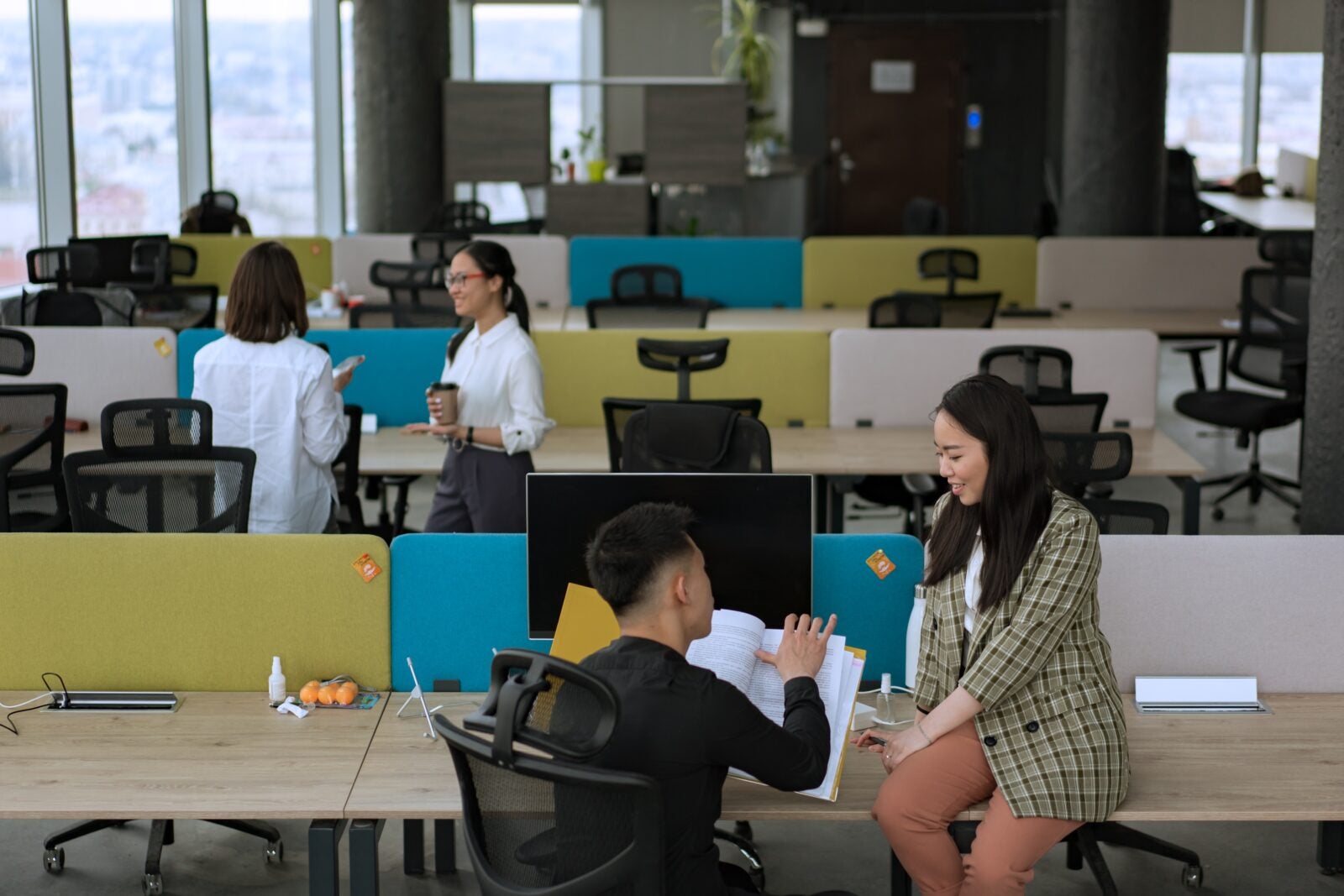 An Office With A Couple Group Of People Who Are Chatting And Generally Chilling. A Woman In Office Attire Is Sitting On A Desk And Talking To Her Male Coworker.