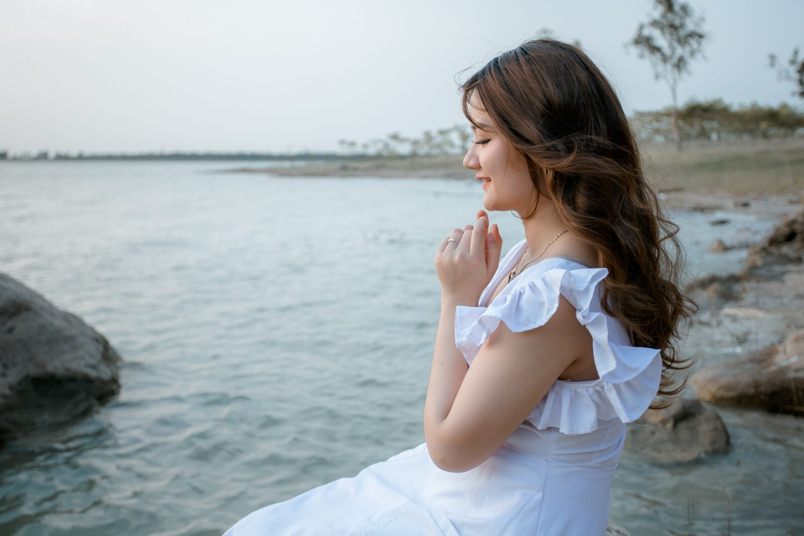 An Asian woman in a white dress sitting by the sea and smiling.