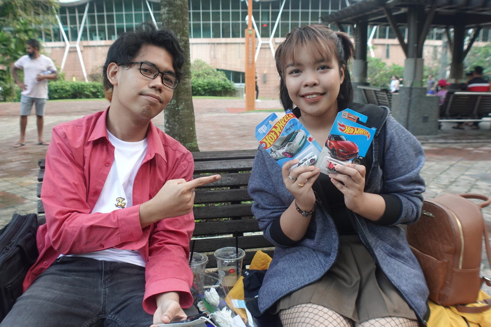A man points to a woman beside him as she poses with a couple of HotWheels that are still in package.