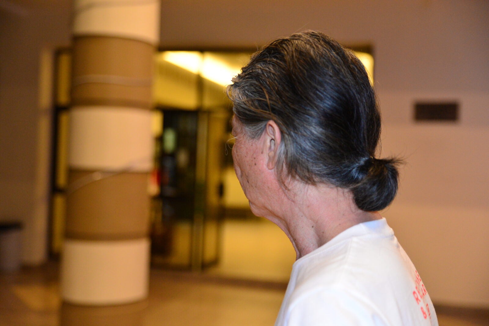 An old man with long hair tied in a bun, wearing a white t-shirt and looking away to hide his face.