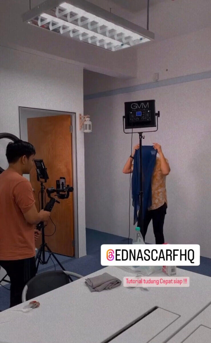 A man is filming himself doing a tutorial of how to wear the hijab that he is selling. He is at a studio and a staff member is recording him with an elaborate looking device and a light stand.