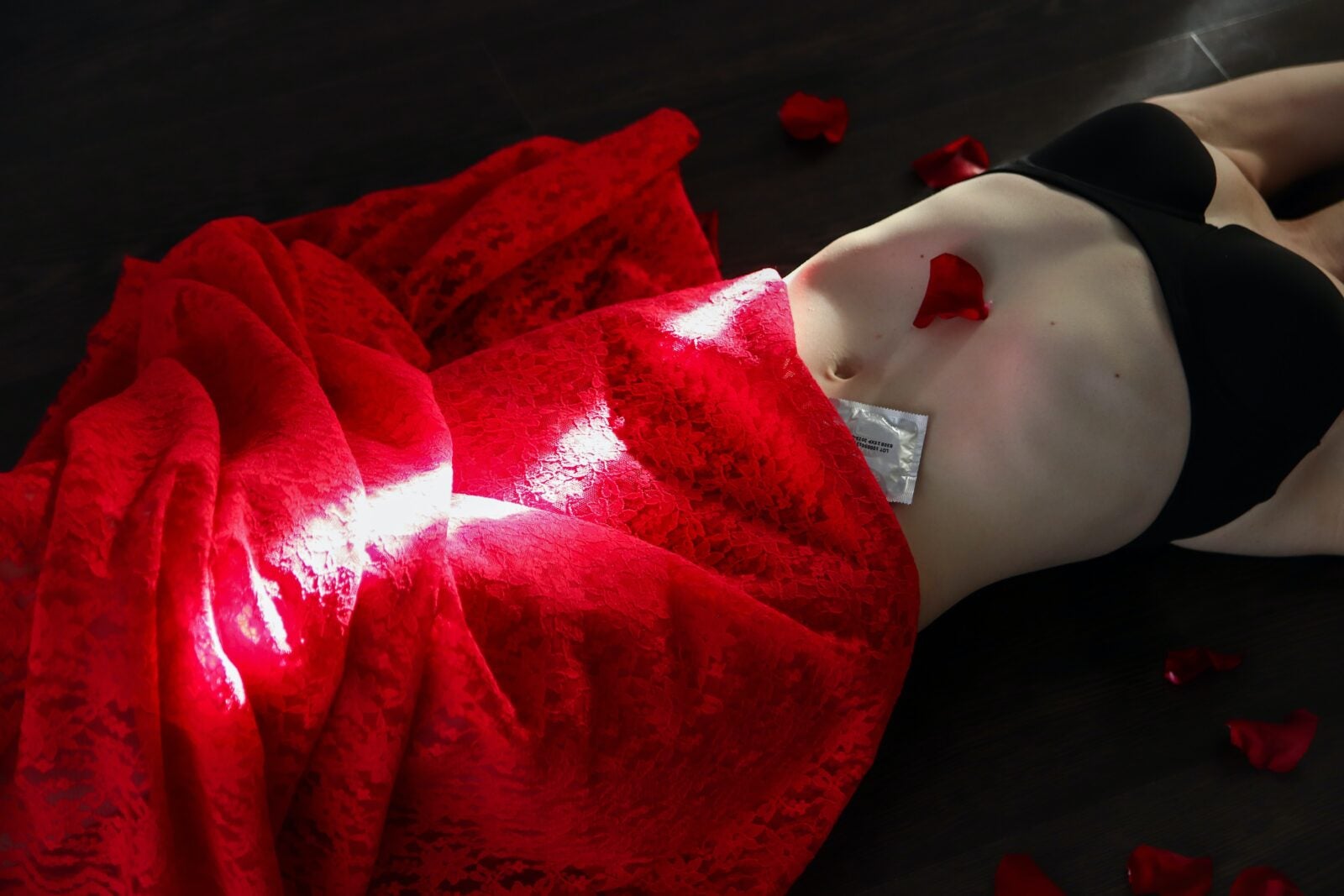 A woman lays down facing the ceiling with a red drape covering the lower half of her body, a rose petal on her abdomen and wearing a black bra. Her hands are stretched upwards and cannot be seen along with her face.