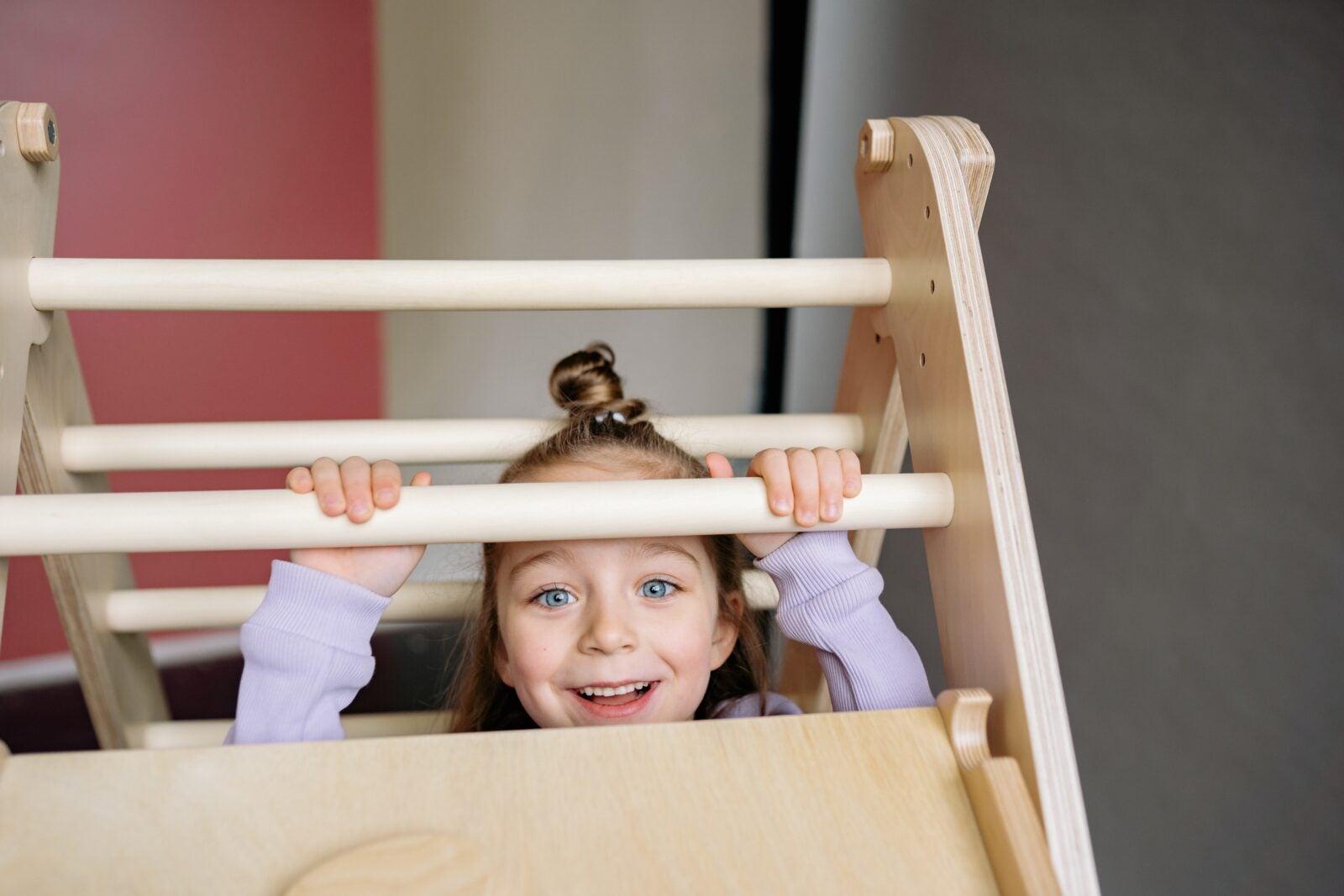 A little girl with blue eyes is smiling while hiding slightly under a wooden construct