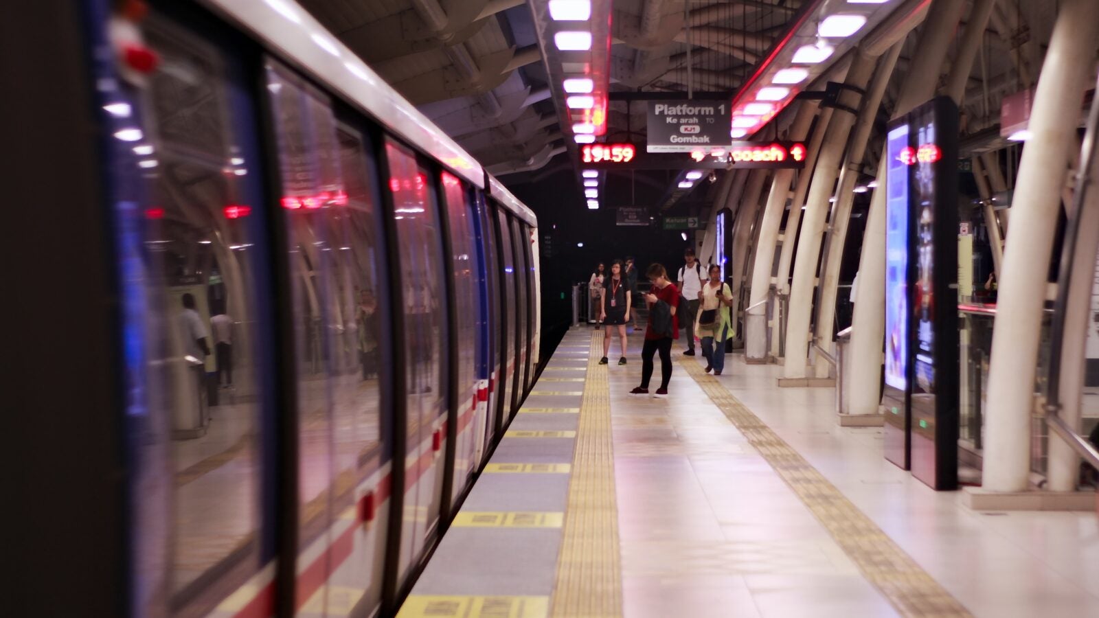 Image shows a wide shot of the waiting area at an LRT station. There are people at the far end, away from the camera. A train is stationed with its door closed at the left side of the image.