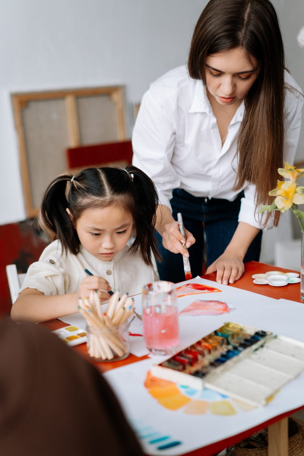 An asian child is doing watercolour art while an older woman stands above her and joins in.