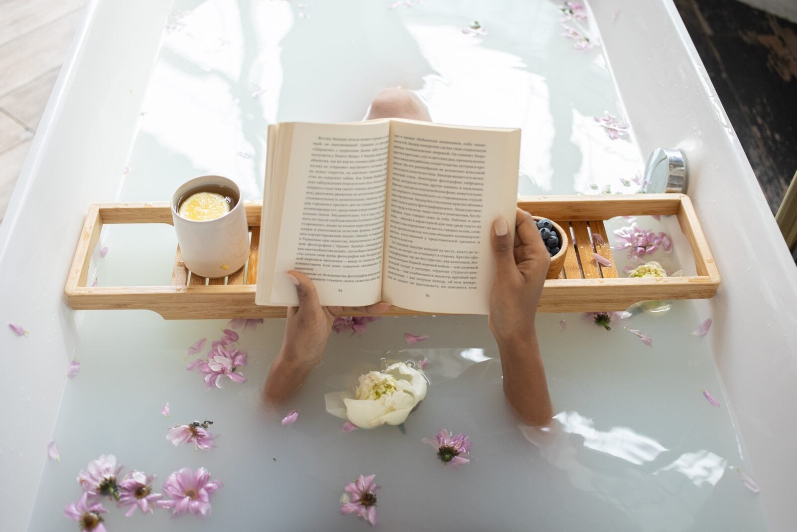 A pair of hands hold a book in a filled bathtub with a wooden platter holding a cup of coffee. There are flowers in the bathwater and the milkiness covers the person's body.