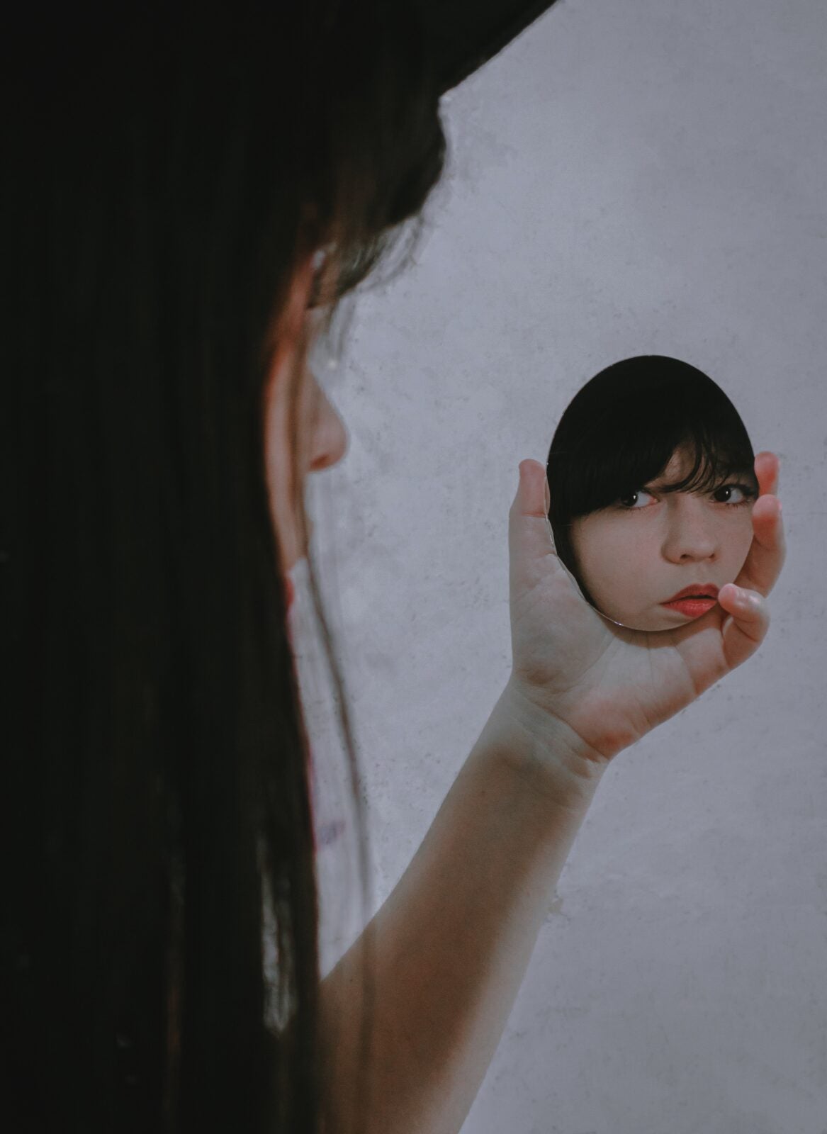 A woman staring at her own reflection through a small, round, hand-held mirror.