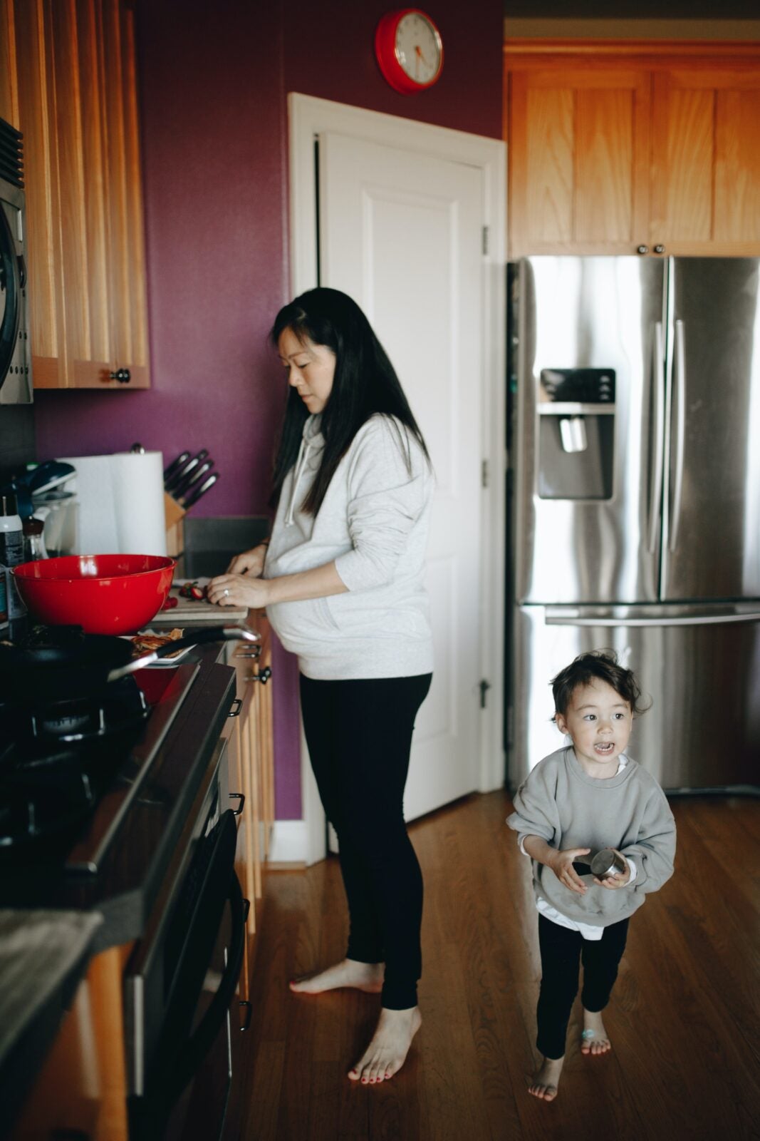 A mother is cooking while her toddler is standing behind her. The toddler has her hands on a toy.