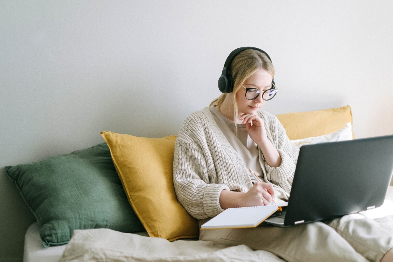 A blonde woman wearing headphones and warm clothes sits on a sofa while using a laptop.