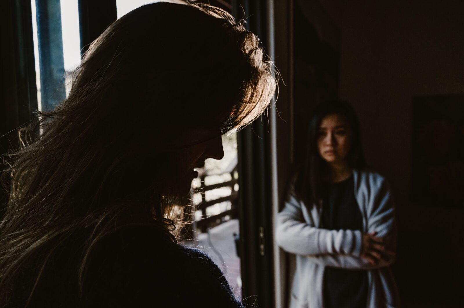 A woman stands by a sliding door and crosses her arms around her chest while looking at someone across from her. The other person is looking down in guilt or shame.