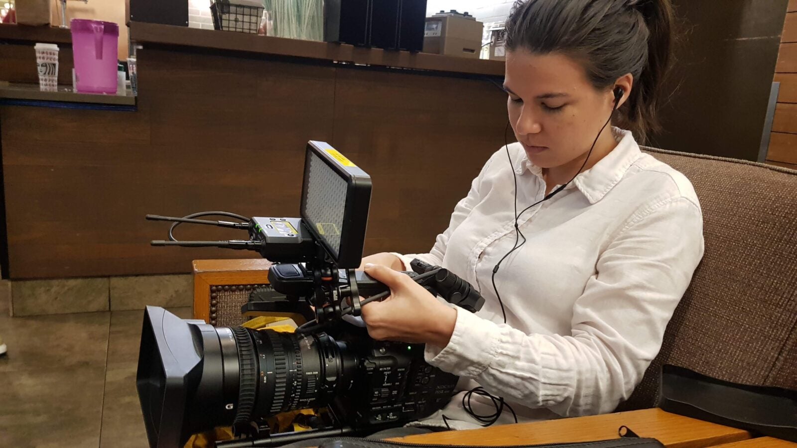 A Swiss-Malaysian Woman sitting on a low chair in a cafe, with earphones on and a huge recording camera on her lap. She is looking down, focusing on the recording camera.
