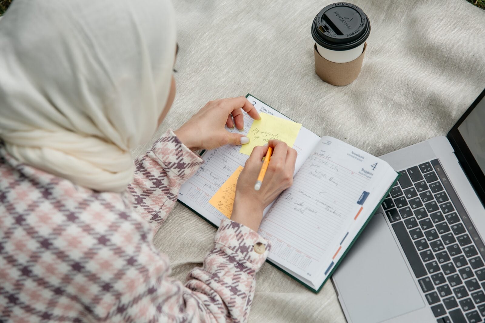 A Muslim Woman wearing the hijab. writing down on a notepad and sitting in front of her laptop.