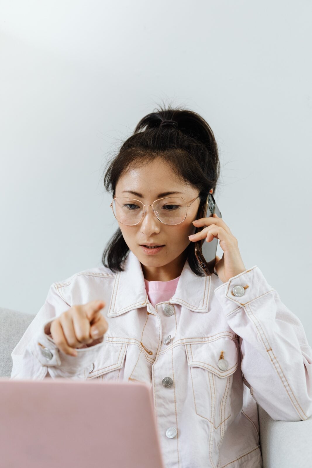 An Asian woman wearing glasses and a jacket using the phone while a pink laptop rests on her lap.