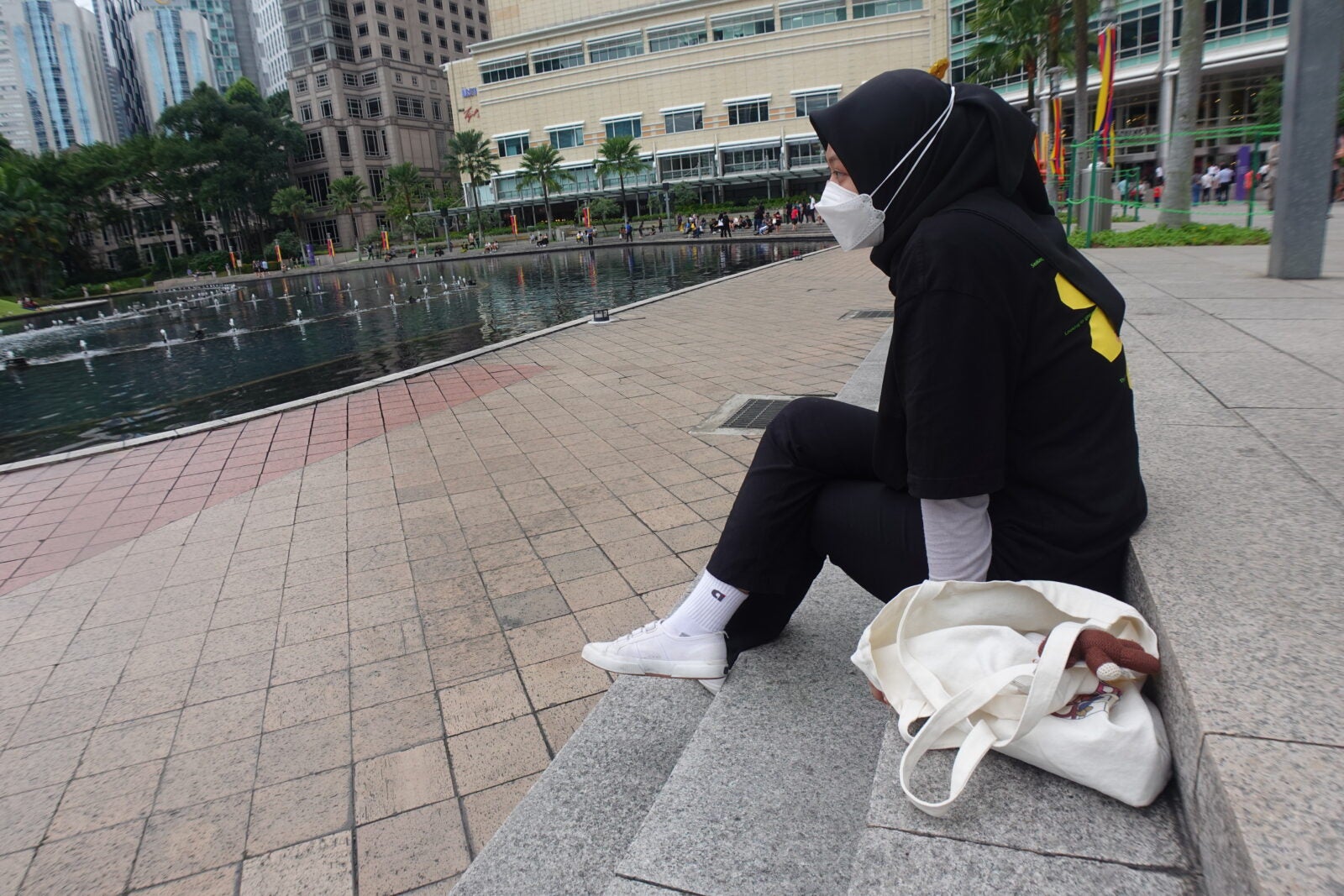 A Malay girl wearing black hijab, and a white face mask, sitting on the stairs at KLCC park.