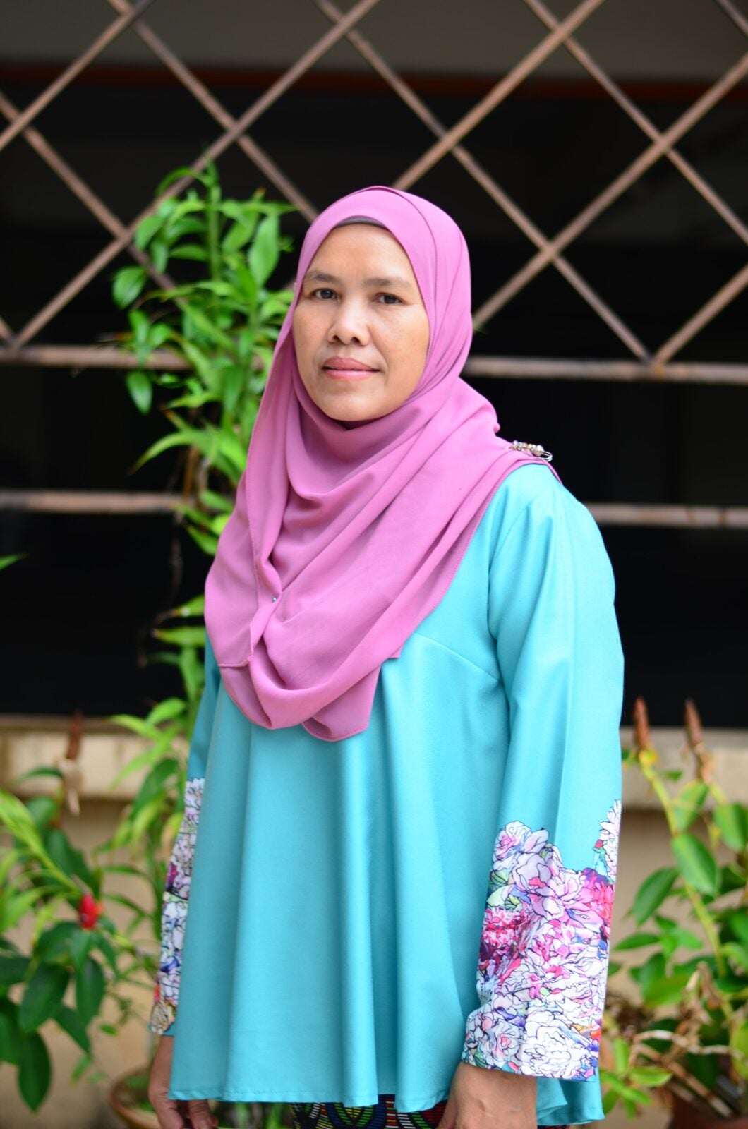 An elderly Malay woman wearing a pink hijab and blue baju kurung.