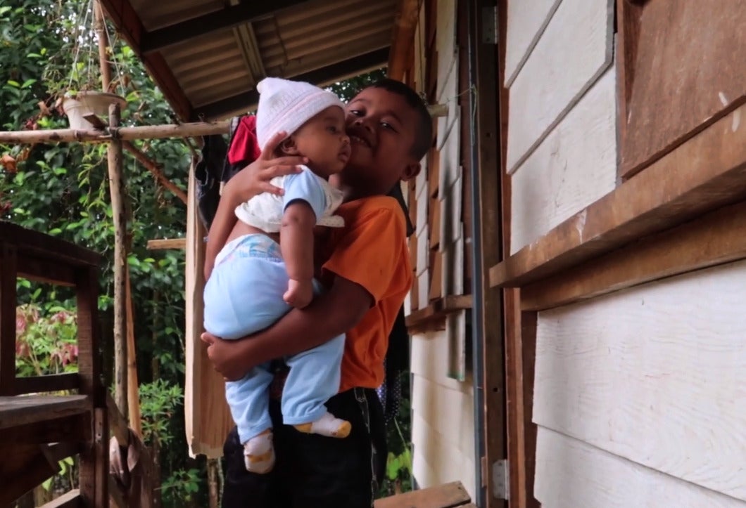 A young Orang Asli boy who is carrying a baby while standing at his Orang Asli family's front porch.
