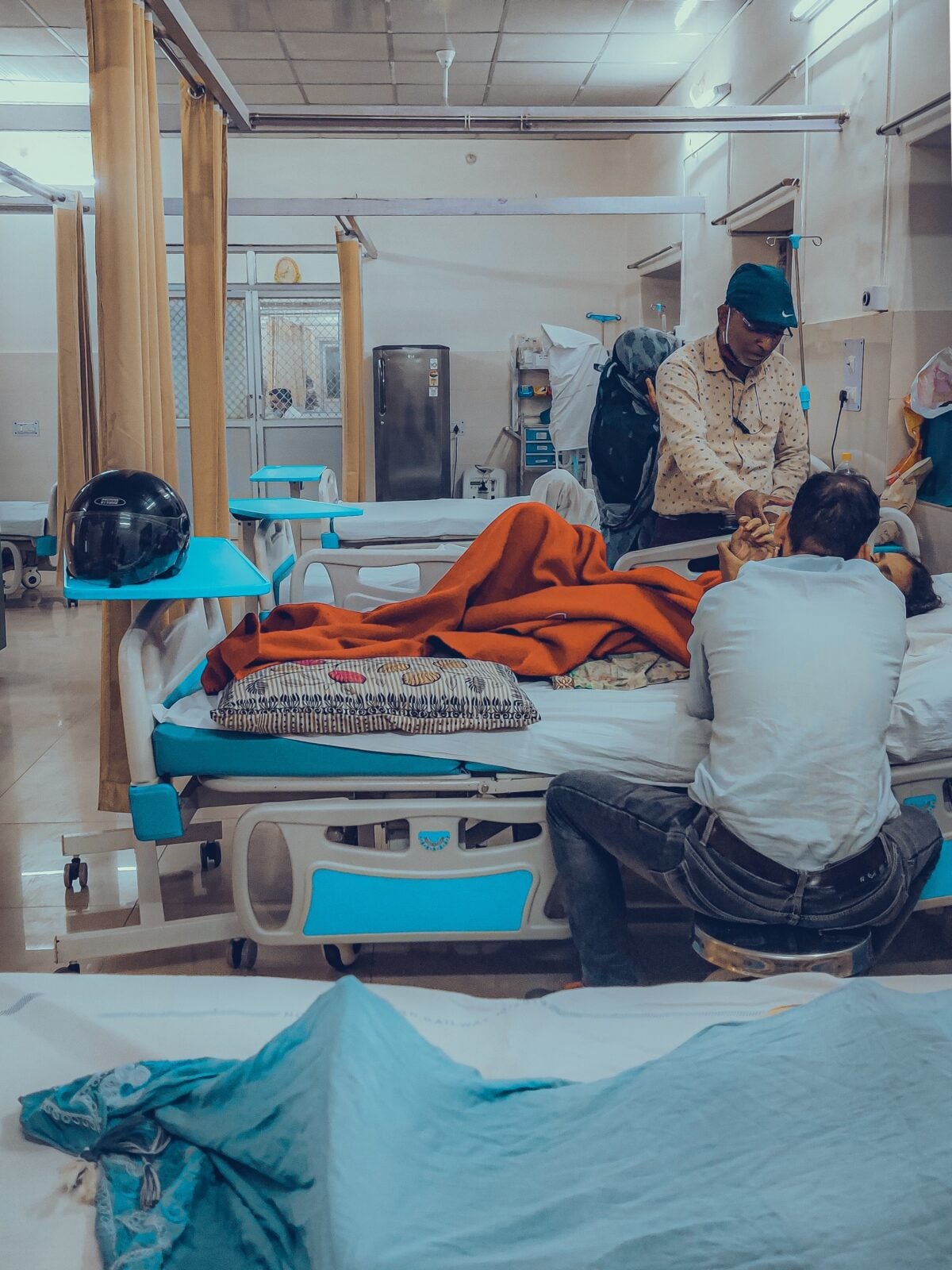 Two men visiting a sick woman who is laying on a hospital bed. 
