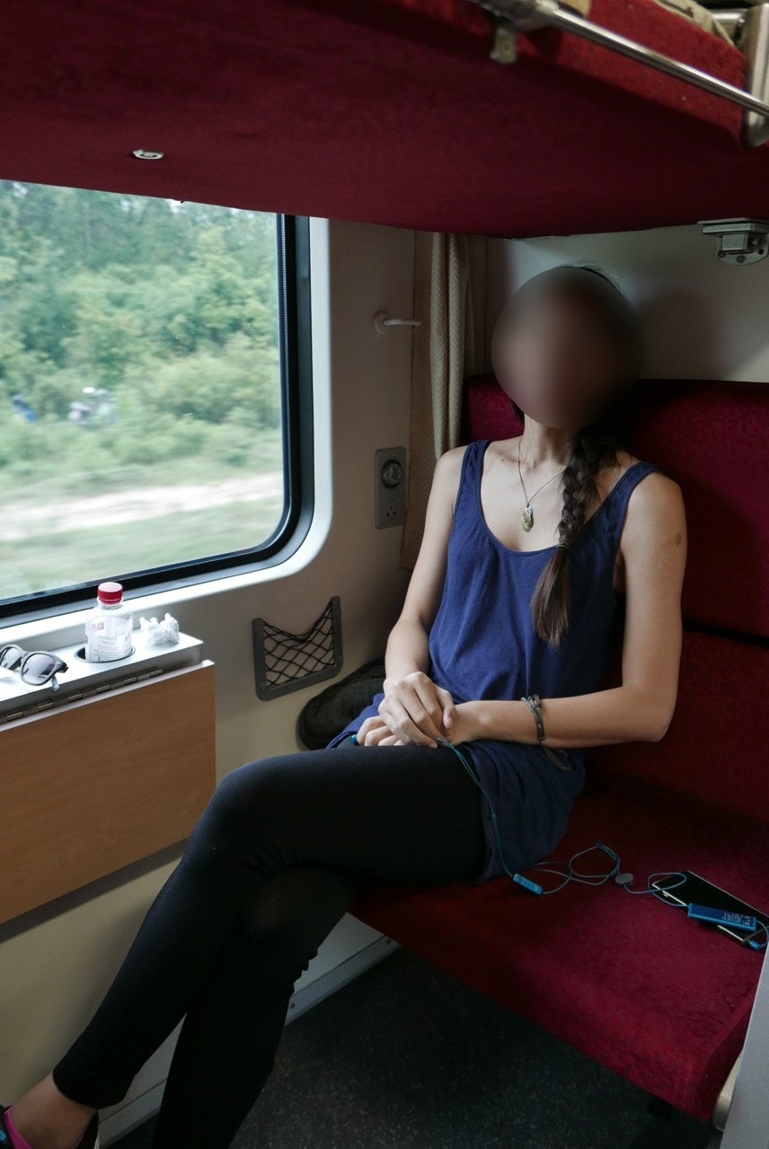 A Transwoman with her face blurred, wearing a tank top and skinny jeans, sitting in a train.