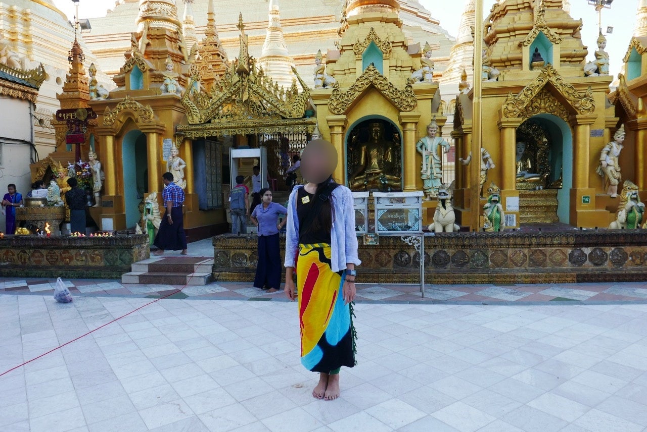 A Transwoman whose face is blurred, wearing a long skirt and standing outside of a temple.