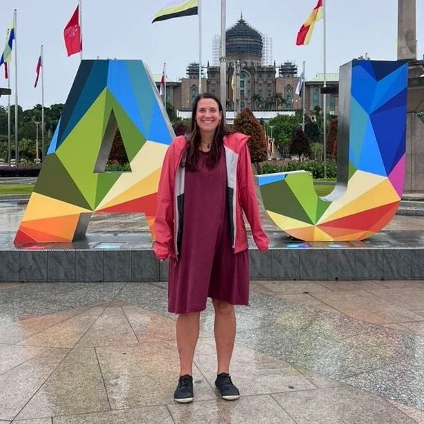 A woman wearing a dress and a windbreaker, standing in front of the Putrajaya sign in Malaysia. 