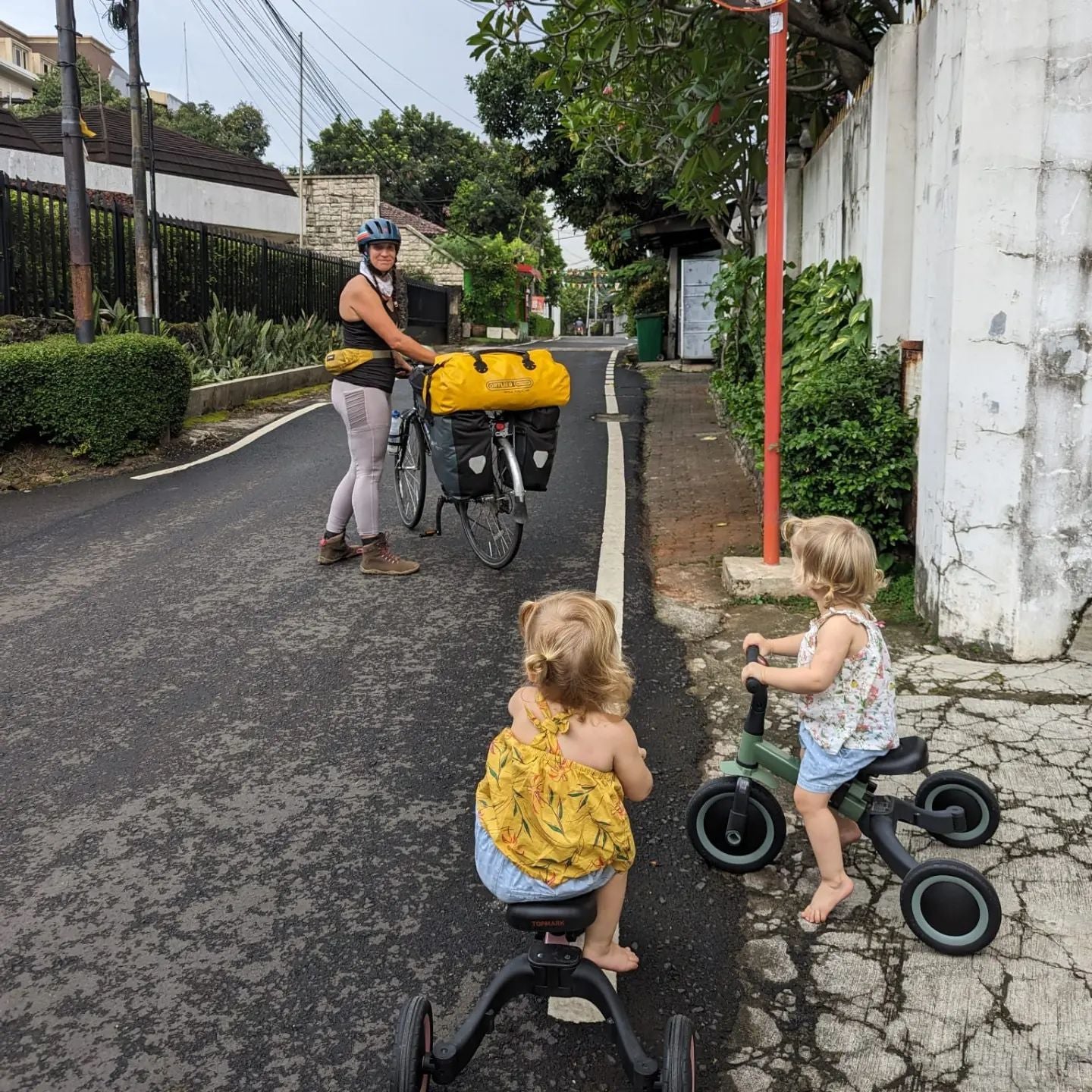 Two children on a tricycle, looking at a woman who is holding her own bicycle which has multiple bags and cases attached to it.