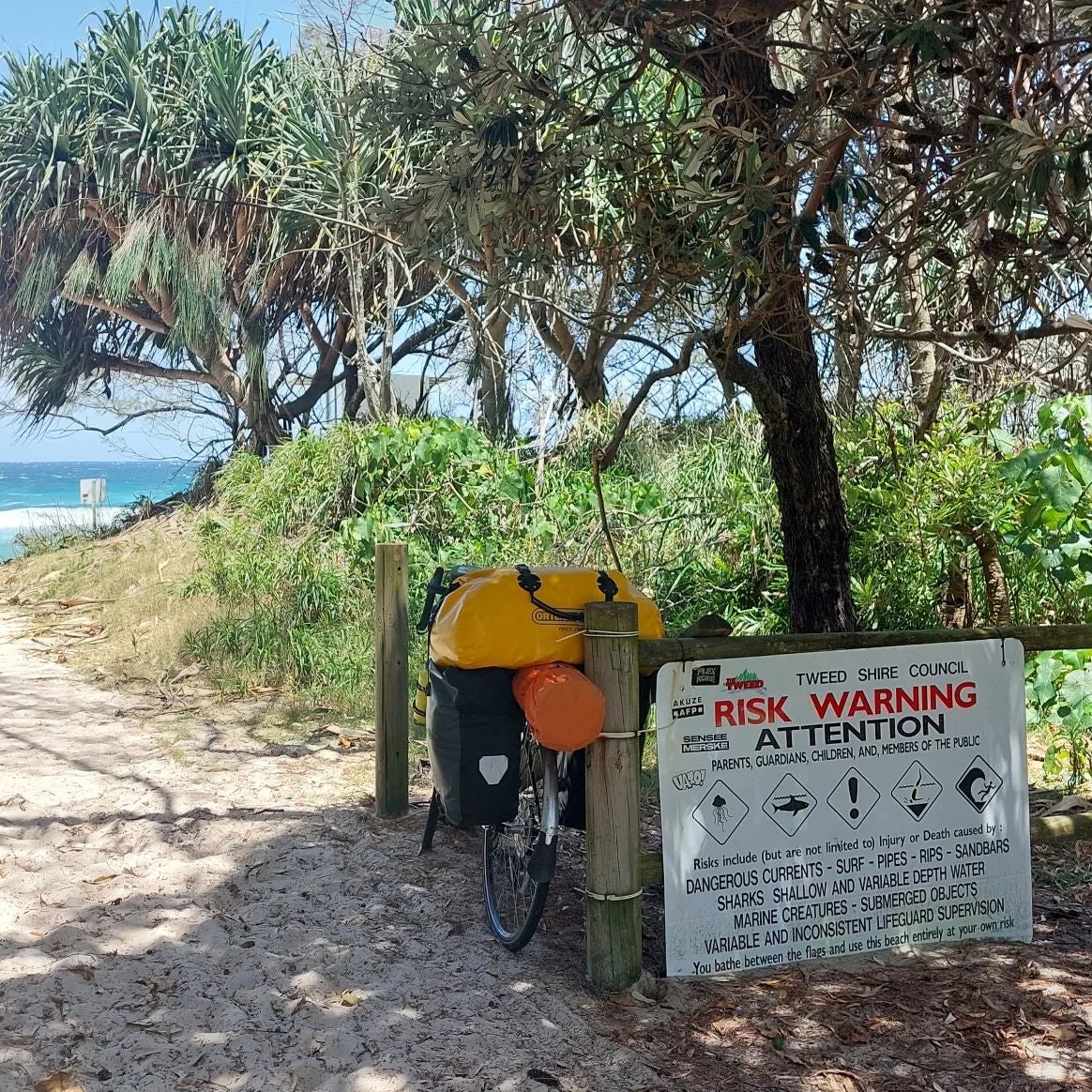 A bicycle with multiple bags attached to its back, resting under a sign that reads: "Risk Warning Attention. Parents, Guardians, Children and Member of the public. Dangerous Currents - Surf - Pipes - Rips - Sandbars - Sharks - Shallow and Variable Depth Water - Marine Creatures - Submerged Objects - Variable and Inconsistent Lifeguard Supervision. Your bathe between the flags and use this beach entirely at your own risk"
