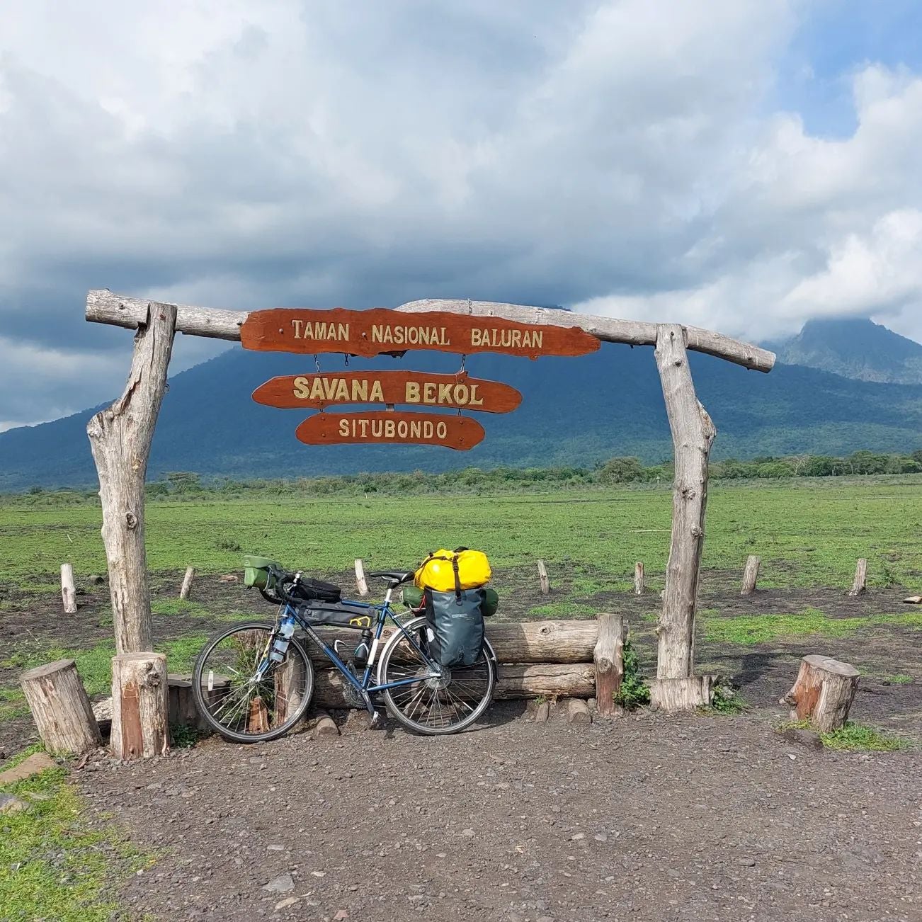 A bicycle with multiple bags attached to its back, resting under a sign that reads: "Taman Nasional Baluran. Savana Bekol Situbondo"
