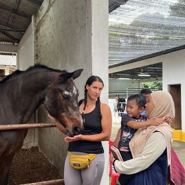 A woman standing in front of a horse, her hands resting under the horse's chin and mouth. A woman wearing the hijab is standing next to her while holding a young boy.