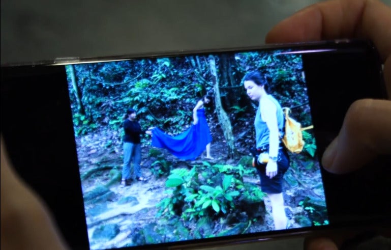 Close up of a phone screen which shows a photo of a Swiss-Malaysian woman in tank top and shorts, looking away from two women who are dressing up in a forest area.