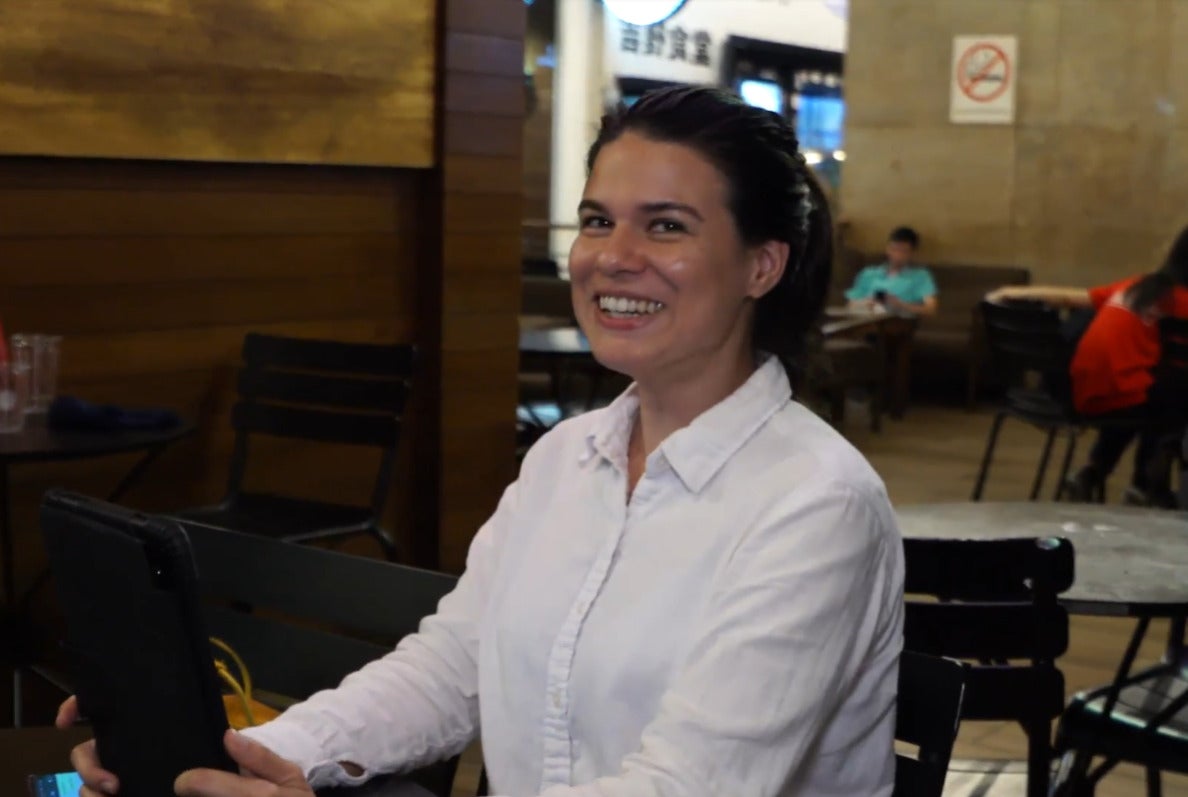 A Swiss Malaysian woman wearing a white shirt. She is using her phone, sitting at a coffee shop and smiling.
