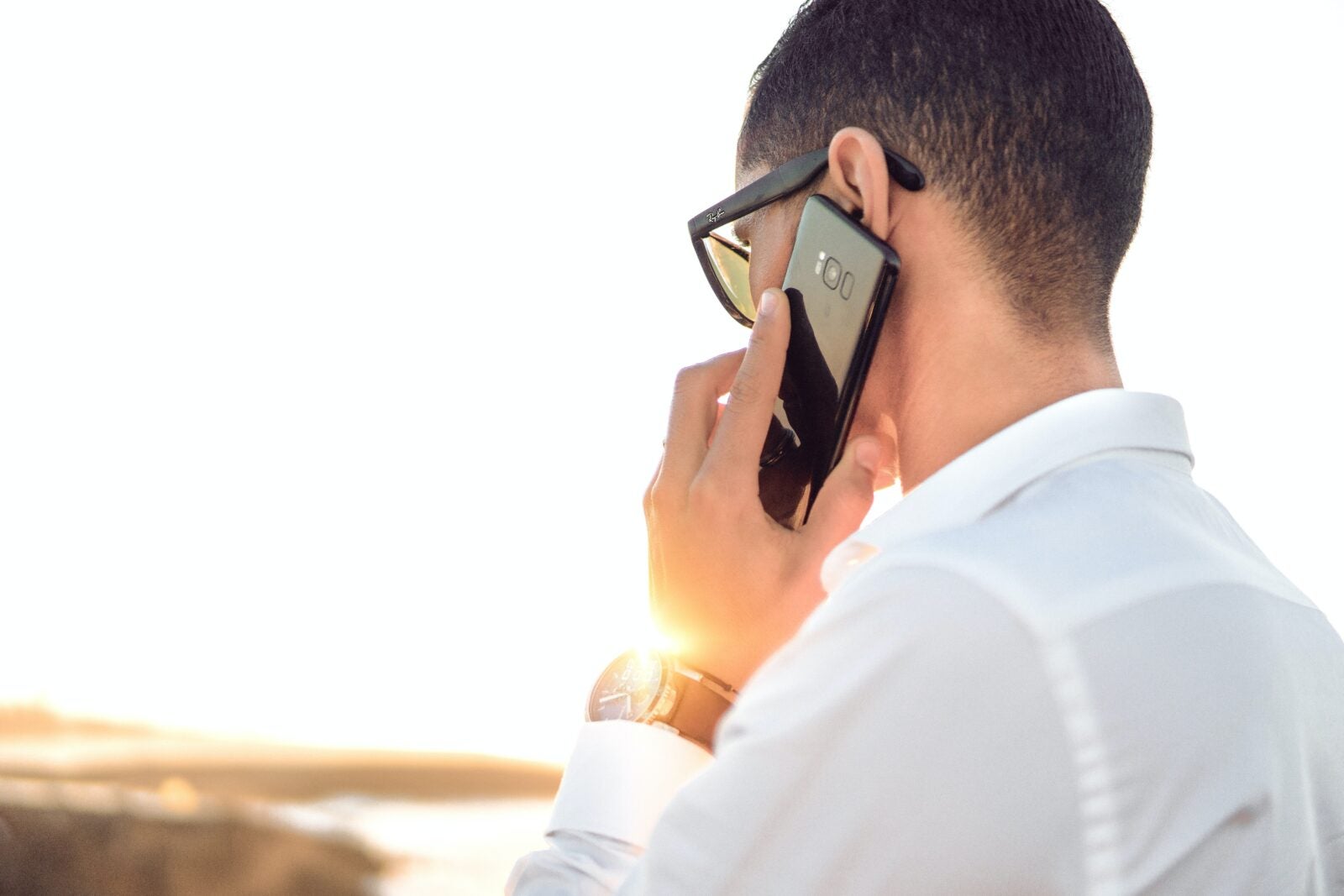 A man wearing sunglasses and a white work shirt, with his phone held to his ear.
